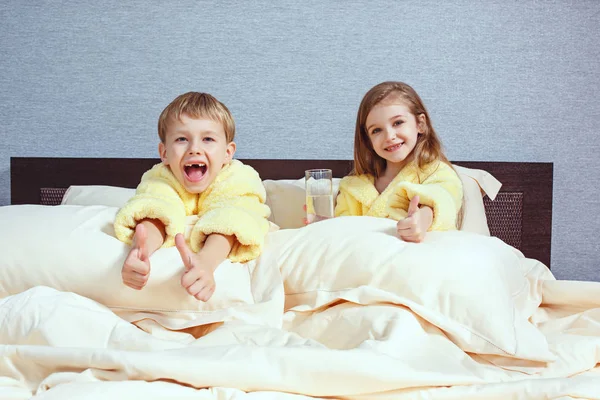 Boldog gyerekek nevetve, fiú és lány puha fürdőköpeny fürdő után játszani fehér ágy — Stock Fotó