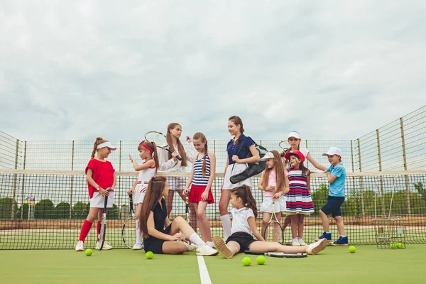 Portrait of group of girls and boy as tennis players holding tennis rackets against green grass of outdoor court. Stylish young teens posing at park. Sport style. Teen and kids fashion concept.