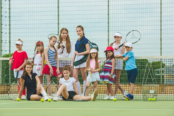 Portrait of group of girls and boy as tennis players holding tennis rackets against green grass of outdoor court. Stylish young teens posing at park. Sport style. Teen and kids fashion concept.