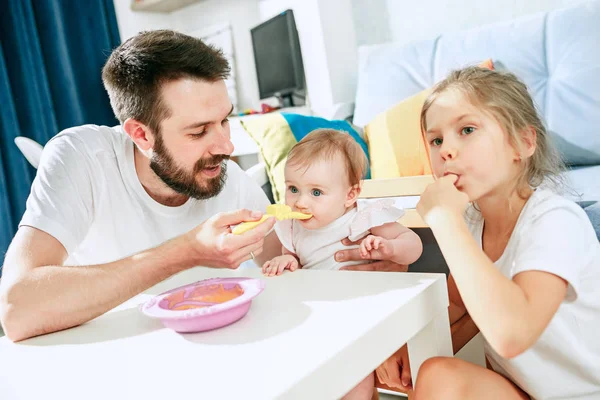 Hombre joven bien parecido desayunando y alimentando a su bebé en casa —  Fotos de Stock