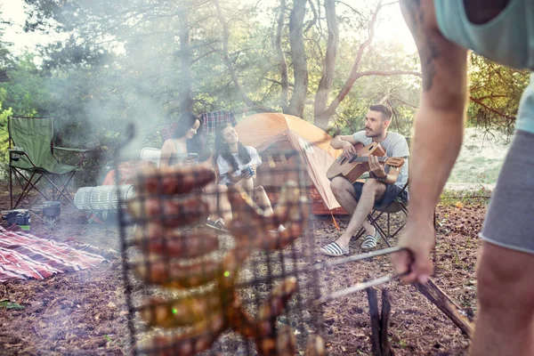 Fiesta, camping de hombres y mujeres grupo en el bosque. Se relajan, cantan una canción y cocinan barbacoa —  Fotos de Stock