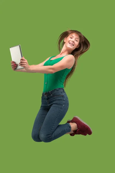 Image of young woman over green background using laptop computer or tablet gadget while jumping.