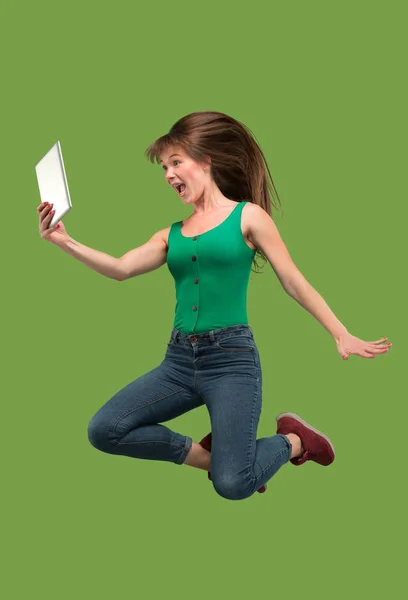 노트북 컴퓨터나 태블릿 가제트를 사용 하 여 점프 하는 동안 녹색 배경 위에 젊은 여자의 이미지. — 스톡 사진