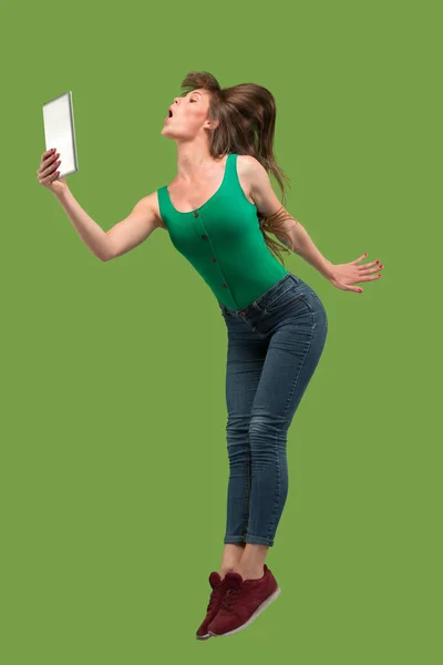 Imagen de mujer joven sobre fondo verde usando computadora portátil o tableta gadget mientras salta . —  Fotos de Stock