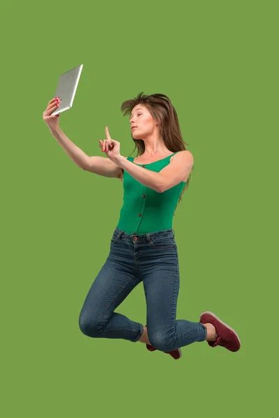 Imagen de mujer joven sobre fondo verde usando computadora portátil o tableta gadget mientras salta . — Foto de Stock