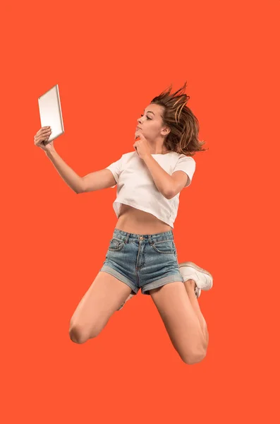 ラップトップ コンピューターやタブレットのガジェットを使用して、ジャンプしながら赤い背景の上の若い女性の画像. — ストック写真