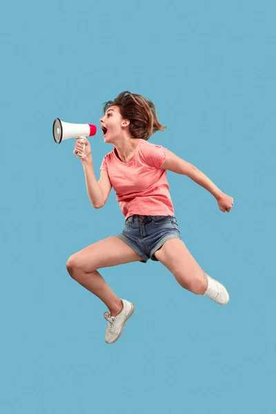 Wanita muda cantik melompat dengan megaphone terisolasi dengan latar belakang biru — Stok Foto