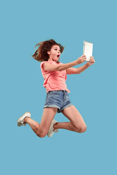 Imagen de mujer joven sobre fondo azul usando computadora portátil o tableta gadget mientras salta . — Foto de Stock