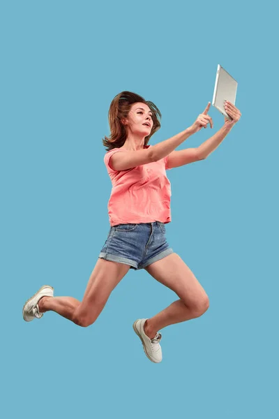 Image of young woman over blue background using laptop computer or tablet gadget while jumping. — Stock Photo, Image