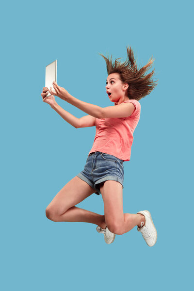 Прыжок молодой женщины на синем фоне студии с помощью ноутбука или планшета гаджет во время прыжка. Бегущая девушка в движении или движении. Концепция человеческих эмоций и выражений лица. Гаджет в современной жизни
