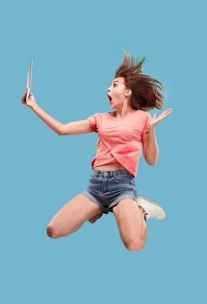 Image of young woman over blue background using laptop computer or tablet gadget while jumping.
