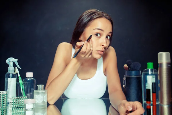 Mulher de beleza aplicando maquiagem. Menina bonita olhando no espelho e aplicando cosméticos com um lápis de olho . — Fotografia de Stock