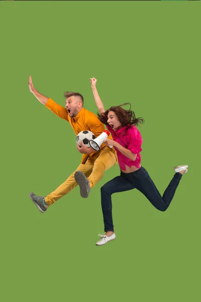 Avance para a vitória.O jovem casal como jogador de futebol saltando e chutando a bola no estúdio em um verde — Fotografia de Stock