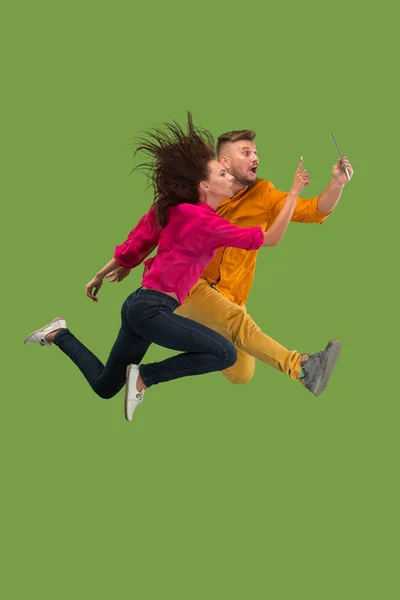 Jump of young couple over green studio background using laptop or tablet gadget while jumping. — Stock Photo, Image