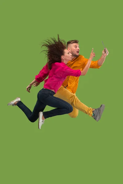Jump of young couple over green studio background using laptop or tablet gadget while jumping.