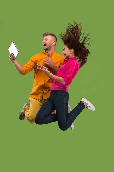 Sauter de jeune couple sur fond vert studio à l'aide d'un ordinateur portable ou tablette gadget tout en sautant . — Photo
