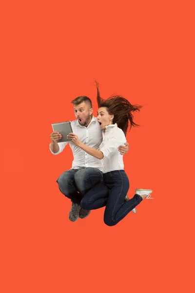 Jump of young couple over red studio background using laptop or tablet gadget while jumping. — Stock Photo, Image