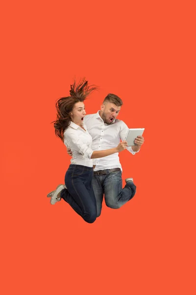 Jump of young couple over red studio background using laptop or tablet gadget while jumping. — Stock Photo, Image