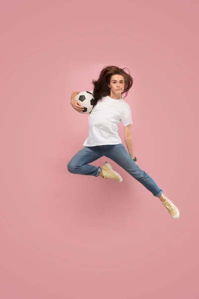 前进的胜利。年轻女子足球运动员跳跃和踢球在工作室粉红色 — 图库照片