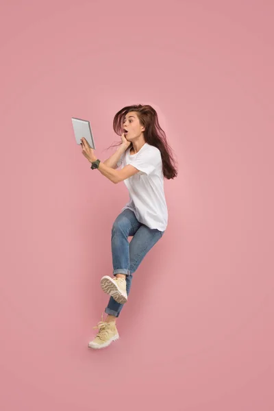 Imagen de mujer joven sobre fondo rosa usando computadora portátil o tableta gadget mientras salta . — Foto de Stock