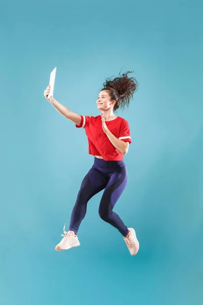 Image of young woman over pink background using laptop computer or tablet gadget while jumping.