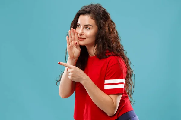 Die junge Frau flüstert ein Geheimnis hinter ihrer Hand vor blauem Hintergrund — Stockfoto