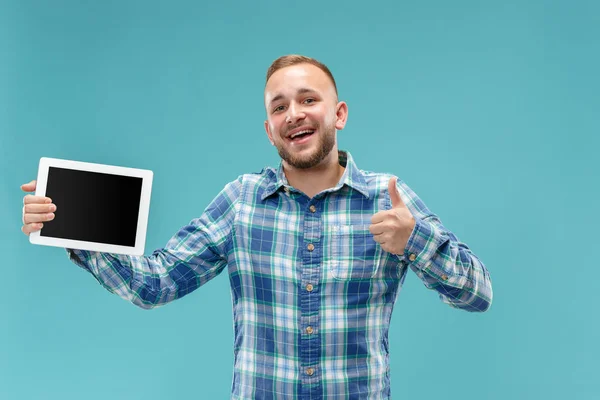 Studio bild av positiva man isolerade på blå bakgrund står i casual kläder håller tablett och visar det tom skärm med lyckligt leende — Stockfoto