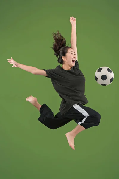 Adelante a la victoria.La joven como futbolista saltando y pateando la pelota en el estudio en verde —  Fotos de Stock