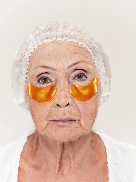 Mujer mayor estudio aislado en la pared blanca concepto de belleza mirando a la cámara haciendo máscaras bajo los ojos —  Fotos de Stock