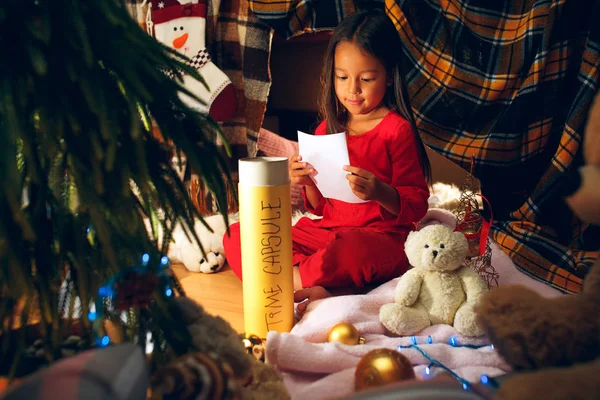 Feliz Navidad y Felices Fiestas. Linda niña escribe la carta a Santa Claus cerca del árbol de Navidad —  Fotos de Stock