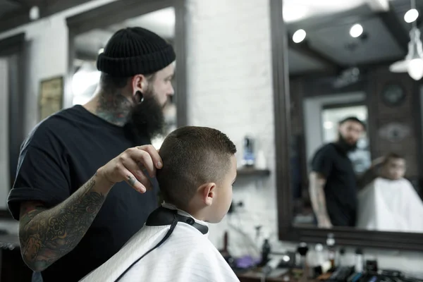 Children hairdresser cutting little boy against a dark background. — Stock Photo, Image