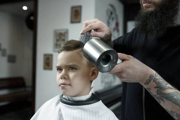 Children hairdresser cutting little boy against a dark background. — Stock Photo, Image