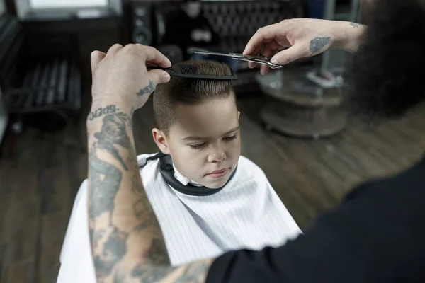 Children hairdresser cutting little boy against a dark background. — Stock Photo, Image