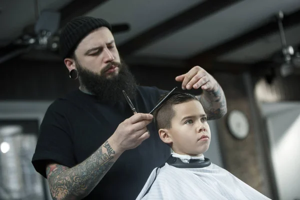 Children hairdresser cutting little boy against a dark background. — Stock Photo, Image