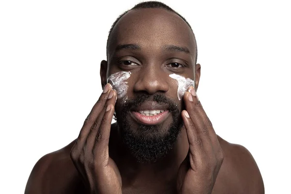 Αφρικανικός-Αμερικανός νεαρός εφαρμογή κρέμας προσώπου κάτω από τα μάτια του σε λευκό φόντο — Φωτογραφία Αρχείου