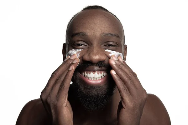 Joven afroamericano aplicando crema facial bajo sus ojos sobre fondo blanco — Foto de Stock