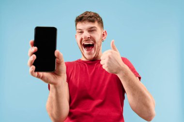 Yakışıklı delikanlı smartphone ekran şok bir sürpriz yüz ile mavi arka plan üzerinde izole gösterilen