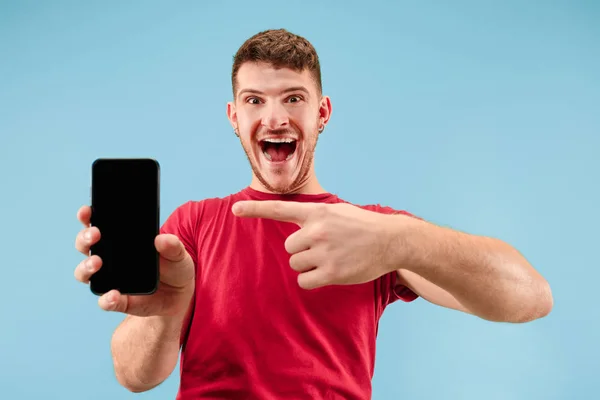 Junger gutaussehender Mann zeigt Smartphone-Bildschirm isoliert auf blauem Hintergrund schockiert mit einem Überraschungsgesicht — Stockfoto