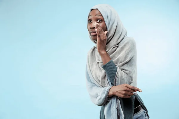 Die junge Afrikanerin flüstert ein Geheimnis hinter ihrer Hand vor blauem Hintergrund — Stockfoto