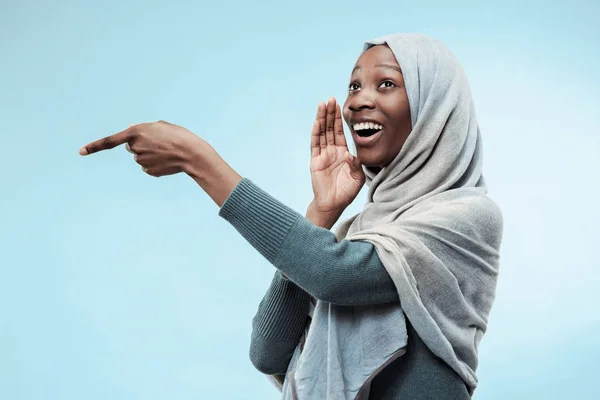 Den unge, afritanske kvinnen som hvisker en hemmelighet bak sin hånd over blå bakgrunn – stockfoto