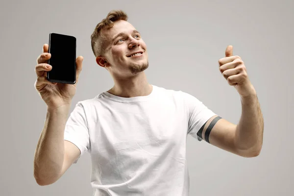 Stilig ung visar smartphone skärmen isolerade på grå bakgrund i chock med en överraskning ansikte — Stockfoto