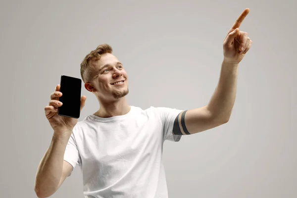 Jovem homem bonito mostrando tela do smartphone isolado no fundo cinza em choque com uma cara surpresa — Fotografia de Stock