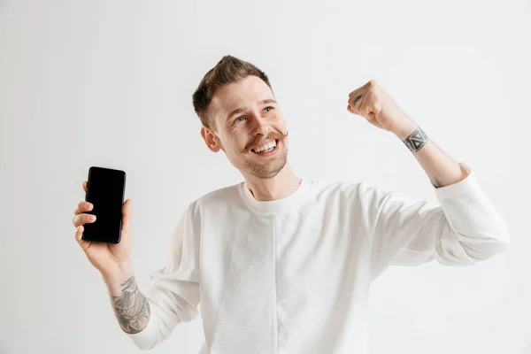 Молодий красивий чоловік показує екран смартфона ізольований на сірому фоні в шоці з несподіваним обличчям — стокове фото