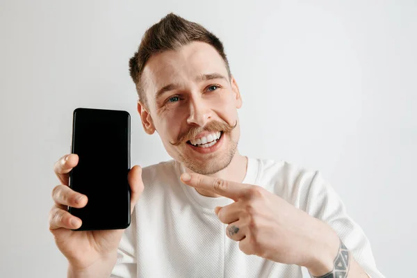 Mladý pohledný muž zobrazení smartphone obrazovky izolované na šedém pozadí v šoku s tváří překvapení — Stock fotografie