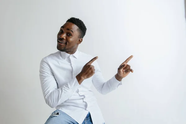 Hombre afroamericano guapo en camisa clásica está sonriendo, mirando a la cámara y apuntando hacia fuera, contra la pared de ladrillo blanco —  Fotos de Stock