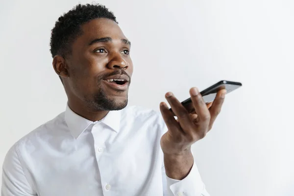 Retrato interior de un atractivo joven africano negro aislado sobre fondo gris, sosteniendo un smartphone en blanco, usando control de voz, sintiéndose feliz y sorprendido. Emociones humanas, concepto de expresión facial — Foto de Stock