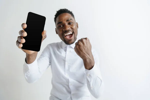 Retrato interior de un atractivo joven africano negro aislado sobre fondo rosa, sosteniendo un smartphone en blanco, sonriendo a la cámara, mostrando la pantalla, sintiéndose feliz y sorprendido. Emociones humanas, faciales — Foto de Stock