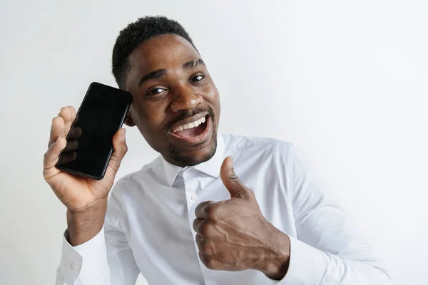 Retrato interior de un atractivo joven africano negro aislado sobre fondo rosa, sosteniendo un smartphone en blanco, sonriendo a la cámara, mostrando la pantalla, sintiéndose feliz y sorprendido. Emociones humanas, faciales — Foto de Stock