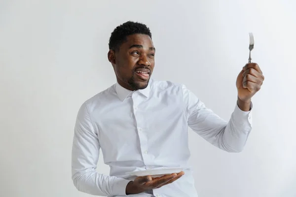 Young african american guy holding empty dish and fork with disgusting facial expression isolated on grey background. Copy space and mock up. Blank template background. — Stock Photo, Image