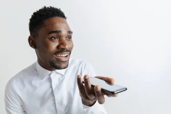 Vonzó fekete-afrikai fiatalember szürke háttér, elszigetelt gazdaság üres smartphone, hangvezérlés használata, érzés, boldog és meglepett beltéri portré. Emberi érzések, arckifejezés koncepció — Stock Fotó